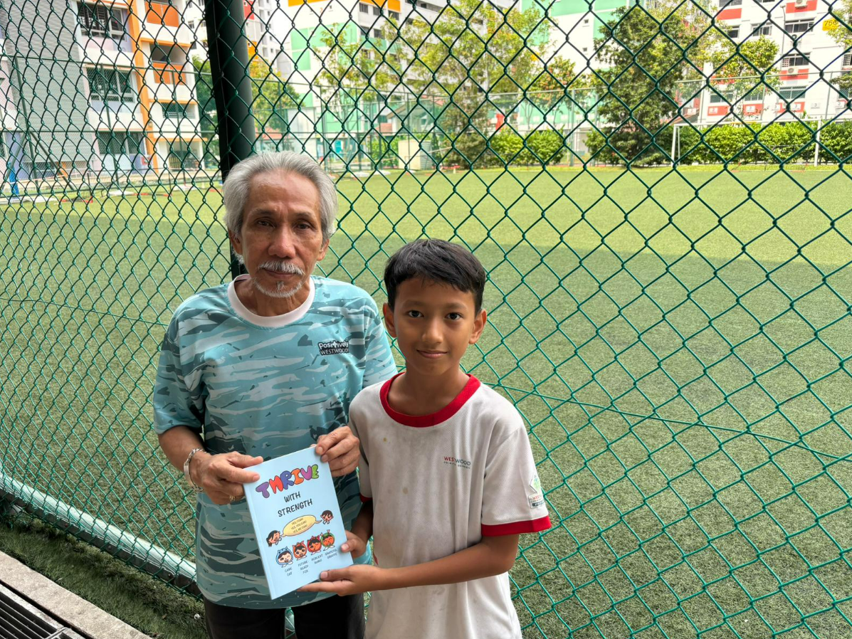 An elderly man and a young boy hold a book titled "Thrive with Strength" by a chain-link fence near a soccer field.